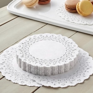 May include: A stack of white paper doilies with a delicate, lacy edge. The doilies are round and sit on a wooden surface. A white tray with macarons is in the background.