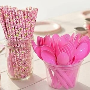 May include: A clear glass jar filled with pink floral patterned paper straws. Next to it, a clear plastic cup overflowing with bright pink plastic spoons. The items are arranged on a white surface, suggesting a party or event setting.