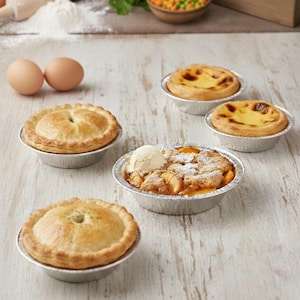 May include: An assortment of baked goods in foil containers, including two savory pies, two custard tarts, and a peach cobbler topped with ice cream. The food is arranged on a light-colored wooden surface.