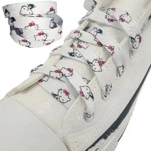 Puede incluir: Zapatillas blancas de lona con cordones blancos que presentan un patrón repetido de un personaje de dibujos animados con lazos rosas y una pajarita azul.