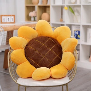Puede incluir: Un cojín de felpa en forma de girasol amarillo con un centro marrón. El cojín está sentado en una silla blanca con un marco de metal dorado.