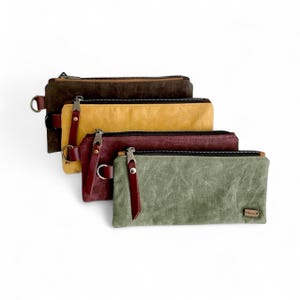 May include: Four rectangular canvas zipper pouches in shades of brown, yellow, burgundy, and green. Each pouch has a leather pull tab and a metal ring. The pouches are arranged diagonally against a white background.