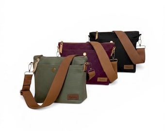 May include: Three canvas messenger bags in olive green, deep purple, and black, each with a brown adjustable shoulder strap. The bags have a rectangular shape with a front pocket and a small logo patch. The bags are displayed against a white background.