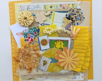 May include: A collection of yellow and brown paper scraps, fabric scraps, and embellishments. The collection includes a yellow crocheted doily, a yellow paper flower, a yellow fabric tassel, a yellow and brown map, and a yellow and brown paper with the word "Enviro" printed on it.