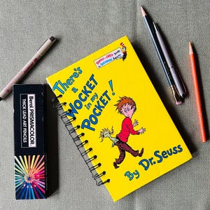 May include: A yellow notebook with a spiral binding features an illustration of a boy with a creature in his pocket. The notebook cover has the title "There's a Wocket in my Pocket!" and is attributed to Dr. Seuss. The notebook is on a green surface with a set of colored pencils and two pens.