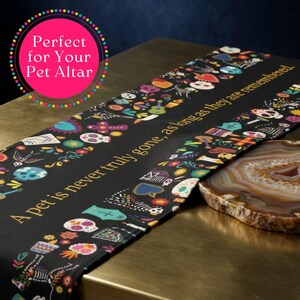 May include: A black table runner featuring a colorful pattern of sugar skulls, flowers, and hearts. The text printed in gold reads: A pet is never truly gone, as long as they are remembered. The fabric has a repeating design of various icons.