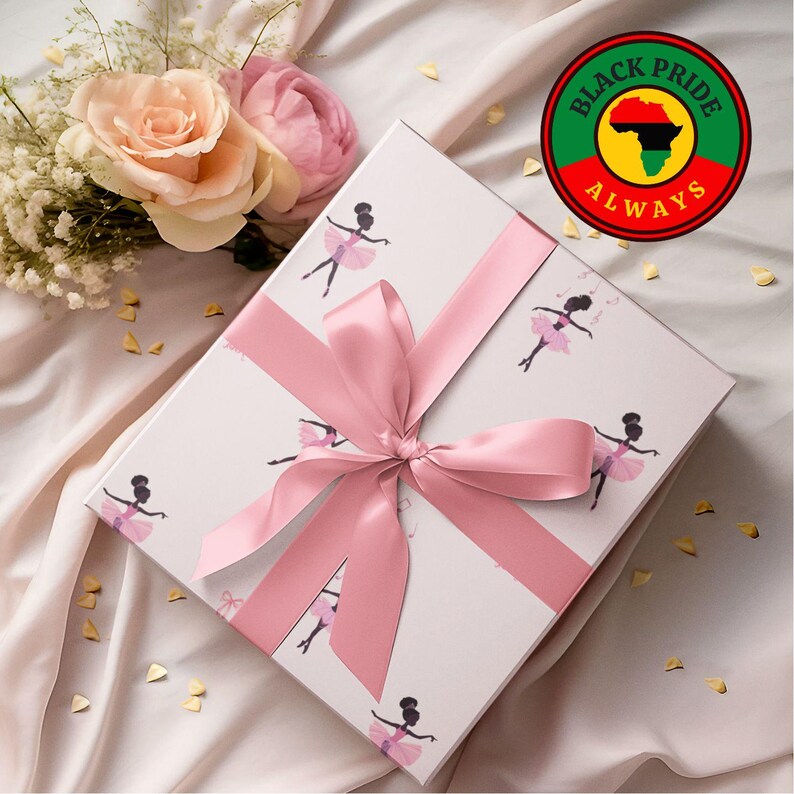 May include: A white gift box with a pink satin ribbon and bow, decorated with illustrations of ballerinas in pink tutus. The box is next to a bouquet of roses and a circular sticker that says "Black Pride Always".