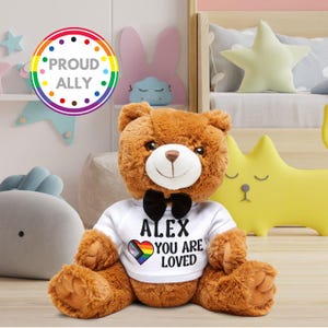 May include: A brown teddy bear wearing a white t-shirt with the text "ALEX YOU ARE LOVED" and a rainbow heart design. The bear has a black bow tie and a smiling face. A "PROUD ALLY" rainbow circle is in the background.