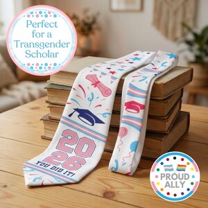 May include: White graduation sash with pink and blue accents, featuring a mortarboard, diploma, stars, and the text "2026 YOU DID IT!". The sash also includes the transgender flag colors and the words "Proud Ally".