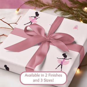 May include: A wrapped gift box with a dusty rose satin ribbon and bow. The white wrapping paper features illustrations of ballerinas in pink tutus. Text on the box reads "Available in 2 Finishes and 3 Sizes!"
