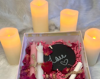 Puede incluir: Una caja de regalo blanca llena de velas rosas y blancas, pétalos de rosa, una pequeña pizarra con la palabra "love" escrita en ella y dos pequeñas botellas de vidrio. La caja está rodeada de cuatro velas blancas.