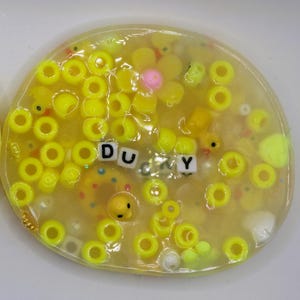 May include: A clear, yellow slime filled with yellow beads, small yellow duck figures, and a pink flower. The word "DU MY" is spelled out with letter beads. The slime is on a white surface.