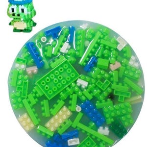 May include: A collection of green, blue, and white building blocks submerged in a clear, gel-like substance. A small, pixelated green figure with a blue hat is positioned to the left of the blocks. The blocks are various shapes and sizes.