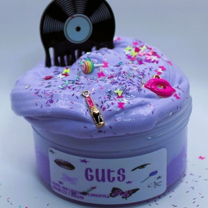 May include: Purple slime in a clear jar with a black vinyl record, a pink lollipop, a gold lipstick charm, and pink and white glitter. The jar has a label that says "Guts" and "Lovespell".