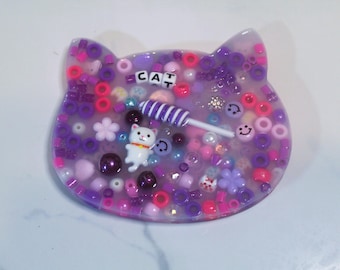 May include: A cat-shaped resin art piece filled with purple, pink, and white beads, flowers, and a white cat figurine. The word "CAT" is spelled out with letter beads. A purple and white striped lollipop is also included.
