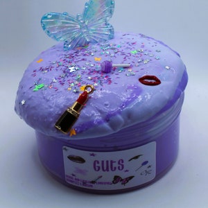 May include: Purple slime with glitter and a butterfly charm, a red lipstick charm, and a lollipop charm. The slime is in a clear jar with a purple label that says "Guts" and has a butterfly, lipstick, and lollipop graphic.