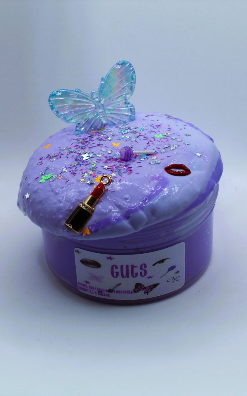 May include: Purple slime with glitter and a butterfly charm, a red lipstick charm, and a lollipop charm. The slime is in a clear jar with a purple label that says "Guts" and has a butterfly, lipstick, and lollipop graphic.