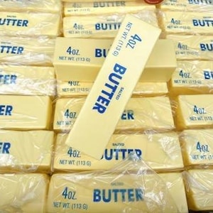 May include: A stack of rectangular sticks of salted butter, each individually wrapped in clear plastic. Each stick is labeled with blue text reading 4oz. NET WT. (113 g) and BUTTER. The sticks are arranged in a grid pattern, with one stick placed diagonally.
