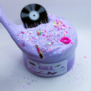 May include: Purple slime with glitter and various charms, including a record, a lipstick, a lollipop, and a pair of lips. The slime is in a clear container with a label that says "Guts".