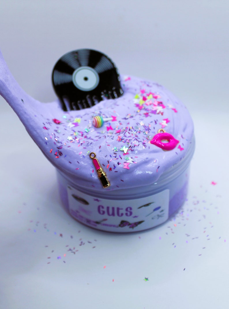 May include: Purple slime with glitter and various charms, including a record, a lipstick, a lollipop, and a pair of lips. The slime is in a clear container with a label that says "Guts".