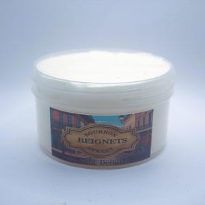 May include: A white plastic container with a clear lid, filled with a white substance. The container has a label with the words "Bourbon Beignets Street" and an illustration of a street scene. The product is a sweet donut.