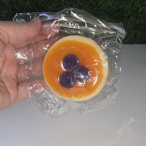May include: A round, off-white soap bar featuring a translucent orange gel topping and three embedded dark purple, spherical accents. The soap is enclosed in a clear, crinkled plastic zip-top bag, showcasing its smooth texture and layered design.