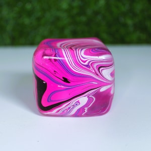 May include: A smooth, cube-shaped object featuring a marbled pattern of vibrant hot pink, deep black, bright white, and subtle purple swirls. The surface has a glossy, reflective finish with rounded edges, creating a fluid, multi-colored design.