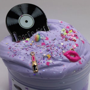 May include: Purple slime with glitter, a black vinyl record, a pink lollipop, a pink lips charm, and a pink lipstick charm.