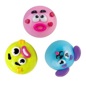May include: Three round, smooth, plastic-like decorative buttons or charms. One is pink with a surprised face and red hearts, one is lime green with a wide-eyed expression and yellow stars, and one is blue with purple ears and a small face.