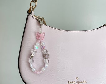 May include: A pink beaded charm with a pink bow and heart-shaped beads is attached to a light pink handbag. The charm features clear, iridescent, and pink beads. The handbag has a silver zipper and the words "kate spade NEW YORK" are printed on the front.