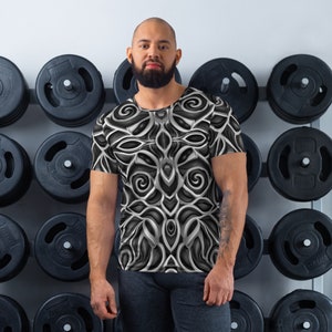May include: A black and white patterned t-shirt with a geometric design. The shirt is worn by a person with a beard in a gym setting.