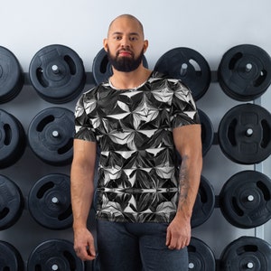 May include: A man wearing a black and white geometric print t-shirt. The shirt has a crew neck and short sleeves. The man is standing in front of a wall with weight plates.