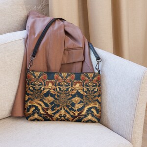 Puede incluir: Un bolso bandolera de color azul oscuro y dorado con estampado y correa negra. El bolso está sobre un sofá de color marrón claro.