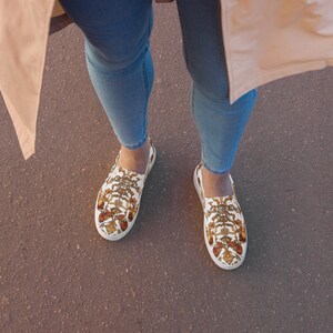 Könnte beinhalten: Ein Paar weiße Canvas-Slipper mit einem gold-braunen Muster. Die Schuhe werden von einer Person getragen, die blaue Jeans und eine beige Jacke trägt.