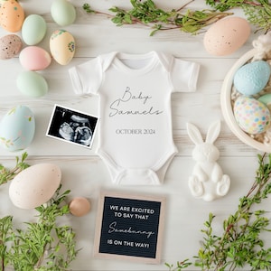 May include: A white onesie with the text "Baby Samaels" and "October 2024" printed on it. A black and white ultrasound image is next to the onesie. A chalkboard sign reads "We are excited to say that Samebunny is on the way!"  Easter eggs and a white bunny figurine are scattered around the onesie.