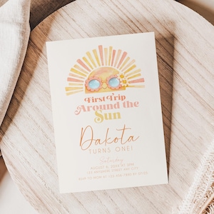 May include: A cream-colored invitation card with the text "First Trip Around the Sun" and "Dakota Turns One!" features a sun illustration with sunglasses. The card is on a wooden plate with a beige linen cloth and dried flowers.