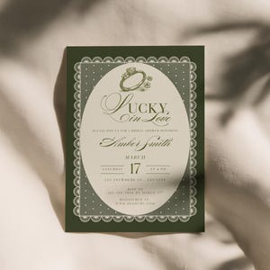 May include: A green invitation card with a white oval in the center, featuring the text "Lucky in Love" and the name "Amber Smith." The card includes details for a bridal shower, with a ring illustration and decorative border.