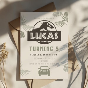 May include: A beige invitation card with a dinosaur graphic and the name "LUCAS" in bold letters. The text "TURNING 5" and event details are also displayed. The card is placed on a beige surface with a brown envelope.