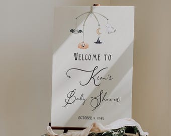 Puede incluir: Un letrero blanco sobre un caballete de madera, dando la bienvenida a los invitados al Baby Shower de Kim. El letrero presenta un móvil caprichoso con una calabaza, un murciélago, un fantasma y una luna. La fecha del evento es el 9 de octubre de 2011.