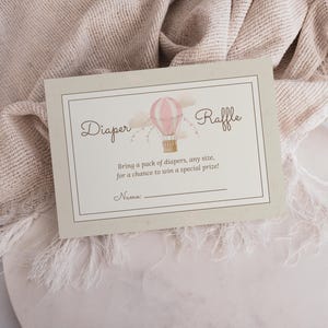 Op de afbeelding: Een rechthoekige kaart met een crèmekleurige achtergrond en een dunne zwarte rand. Het bevat een aquarelillustratie van een roze en witte heteluchtballon. De tekst luidt: Diaper Raffle, Bring a pack of diapers, any size, for a chance to win a special prize!, Name:.