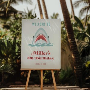 May include: A wooden easel displays a sign with the words "Welcome to" above a cartoon shark emerging from waves. Below, in a decorative font, it reads "Miller's 5th Birthday" and the date. The sign is set against a backdrop of lush greenery.