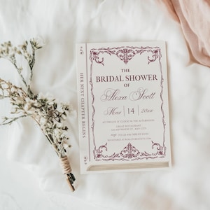 May include: A bridal shower invitation designed to look like a book. The invitation is white with burgundy text and decorative borders. The text reads "The Bridal Shower of Alexa Scott" with the date and time. A small bouquet of dried flowers rests beside the invitation.