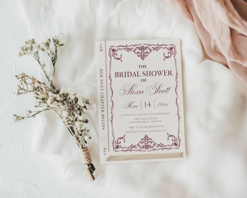 May include: A bridal shower invitation designed to look like a book. The invitation is white with burgundy text and decorative borders. The text reads "The Bridal Shower of Alexa Scott" with the date and time. A small bouquet of dried flowers rests beside the invitation.