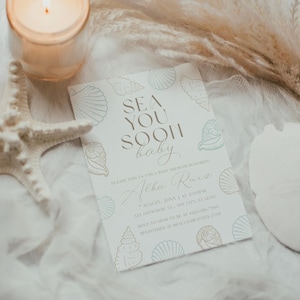 Może przedstawiać: Zaproszenie na baby shower z napisem "SEA YOU SOON baby" i ilustracjami muszli. Zaproszenie otoczone jest rozgwiazdą, świecą i suszonymi roślinami, tworząc estetykę w stylu plażowym.