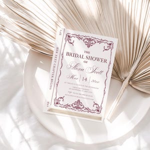May include: A bridal shower invitation designed to look like a book, with the text "The Bridal Shower of Alexa Scott" in elegant script. The invitation is white with burgundy accents and is set on a white plate with a dried palm leaf.