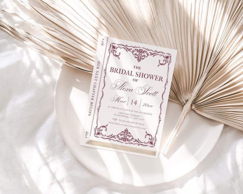 May include: A bridal shower invitation designed to look like a book, with the text "The Bridal Shower of Alexa Scott" in elegant script. The invitation is white with burgundy accents and is set on a white plate with a dried palm leaf.