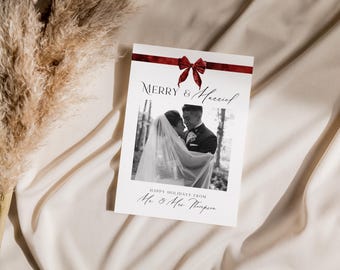 May include: A black and white photograph of a couple in a woodland setting. The photo is on a white card with a red ribbon and bow. The text "Merry & Married" is written in black script on the card. The text "Happy Holidays from Mr. & Mrs. Thompson" is written in black script at the bottom of the card.