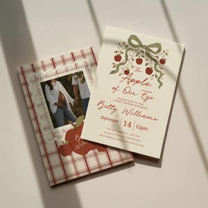 May include: Two rectangular paper cards featuring a cream background. One card displays a red and white plaid pattern with a lace border and a photo of a pregnant person. The other card features a green bow, red apples, and the text The Apple of Our Eye.