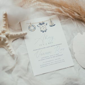 Nautical-themed baby shower invitation reading “Ahoy It’s a Boy,” featuring watercolor illustrations of a sailor onesie, sailboat, and lifesaver hanging on a rope. Styled elegantly on a soft fabric background, accented with natural starfish, shells, and pampas grass, perfect for coastal-themed or seaside baby showers.