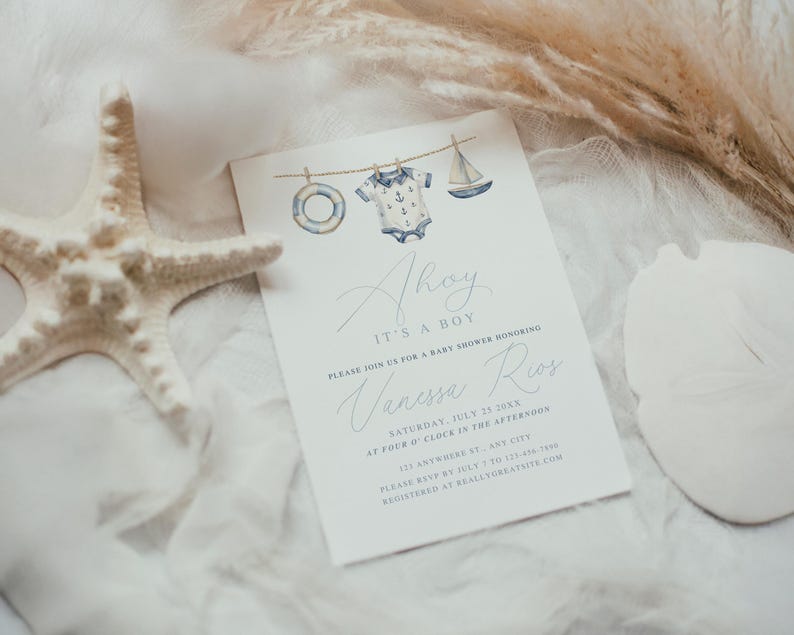 Nautical-themed baby shower invitation reading “Ahoy It’s a Boy,” featuring watercolor illustrations of a sailor onesie, sailboat, and lifesaver hanging on a rope. Styled elegantly on a soft fabric background, accented with natural starfish, shells, and pampas grass, perfect for coastal-themed or seaside baby showers.